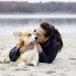A woman lays on the sand at a beach near a lake, hugging and kissing a corgi laying next to her.