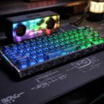 A blue-lit mechanical keyboard and a rainbow-colored speaker on a desk.