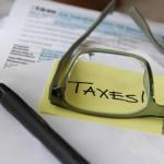 A low-angle view of tax documents and the word 'Taxes' written on a yellow sticky note viewed through eyeglasses.
