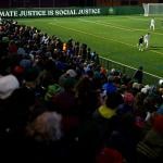 A view from the stands of the match with fans visible in the shadows on the bottom left and the green soccer pitch on the top right, with the words "Climate justice is social justice," displayed behind the goal during the  between the Vermont Green FC and Lexington SC during the 2024 U.S. Open Cup at Virtue Field on March 19, 2024 in Burlington, Vermont. 