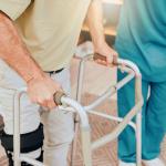 A senior man using a walker during a physiotherapy session assisted by a caregiver.