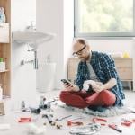 A man watching something on his smartphone while repairing a sink at home.