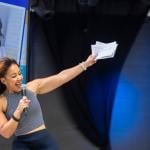 Julia Collins who is the founder and CEO of Planet FWD, speaks onstage with one hand holding a microphone and the other arm raised, her portrait is visible on a screen in the background.