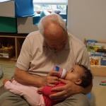 Teacher Herb Hickey holds a baby while she drinks from a bottle.