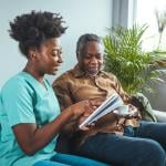 A black mid-adult nurse attentively aids an elderly black male with his reading material.