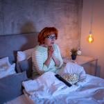 A senior woman watching a scary movie on the TV from bed.