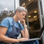 A female small business owner by the entrance of her store reviewing information on a laptop.
