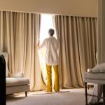 A senior woman standing by the large window of a cozy living room.
