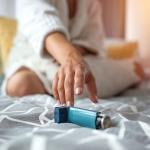 Woman sitting on a bed reaching for her inhaler.
