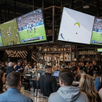 People surround a bar where multiple TVs stream a football match.