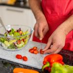 A person slicing cherry tomatoes to a greens salad in the kitchen.