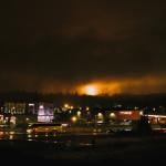 A large flame from a gas flare in Kitimat is visible at night, with the city streets visible in the foreground.