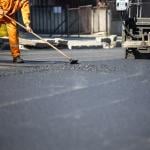 A construction worker in an orange uniform spreads freshly poured asphalt on a road surface.