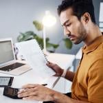 A finance analyst reviewing financial documents using a calculator and a work laptop in an office.
