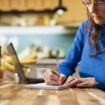 An elderly person writing a grocery list in a kitchen table.