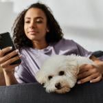 A woman relaxing on the couch while scrolling through her phone and petting her dog.