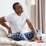 A man sitting on a bed experiencing lower back pain.
