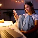 A young woman sitting at a desk in her bedroom and browsing her phone at night.