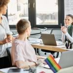 Business partners collaborating on a project and a rainbow flag seen from a desk indicating it as a LGBTQ+ inclusive workplace. 