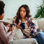 Two female friends having a discussion at home while drinking coffee.
