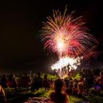 Crowds of people sit in lawn chairs and blankets to enjoy the colorful fourth of July independence day celebratory fireworks exploding in the black night sky.