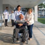 A female doctor assisting her patient in pushing his wheelchair along with a companion.