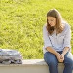 An upset teenage girl sitting on a curb and staring at her backpack for school.