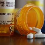 A close up of two orange bottles on a table, one standing upright with its prescription label for oxycodone visible, and the other on its side with the top off and a few white pills spilling out.