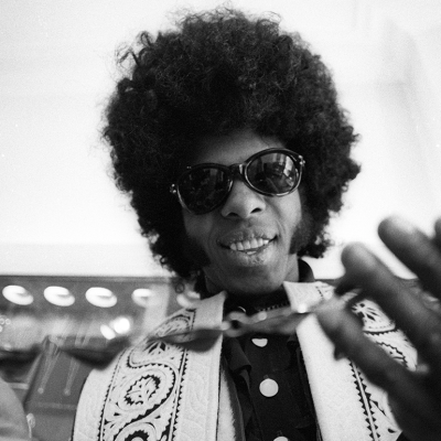 Sly Stone wearing sunglasses, holding a necklace.