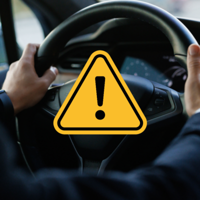 Person’s hands on steering wheel of a Tesla with yellow caution symbol overlaid in the center of the photo.