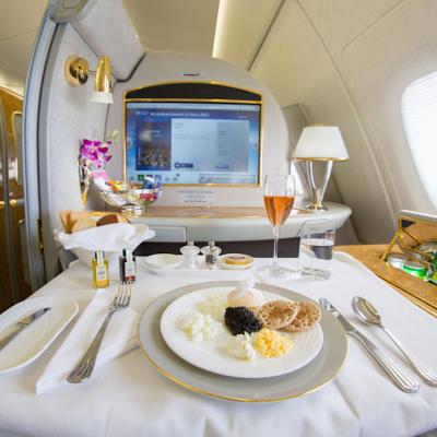 Emirates airlines first class suite with a large screen, a mini bar, and a table set with a meal and a drink in a glass flute.
