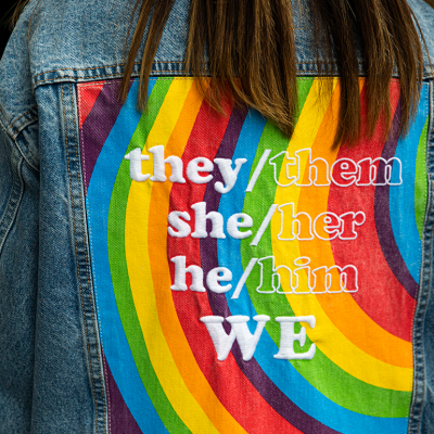 View of the back of a person wearing a denim jacket with gender neutral pronouns over a rainbow print.