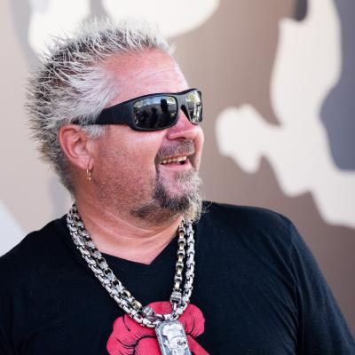 Restauranteur Guy Fieri smiles in sunglasses, chains, and a black T-shirt at the 2022 Stagecoach Festival in Indio, California.