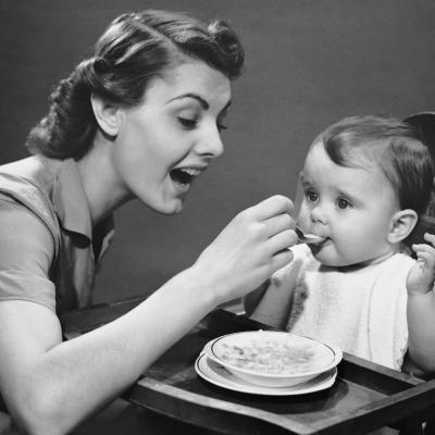 Woman feeding baby in a high chair in the 1950s.