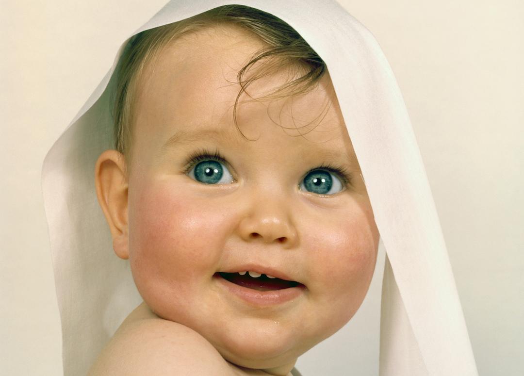Baby with big blue eyes smiling with a long piece of toilet paper draped over her head in the 1960s.