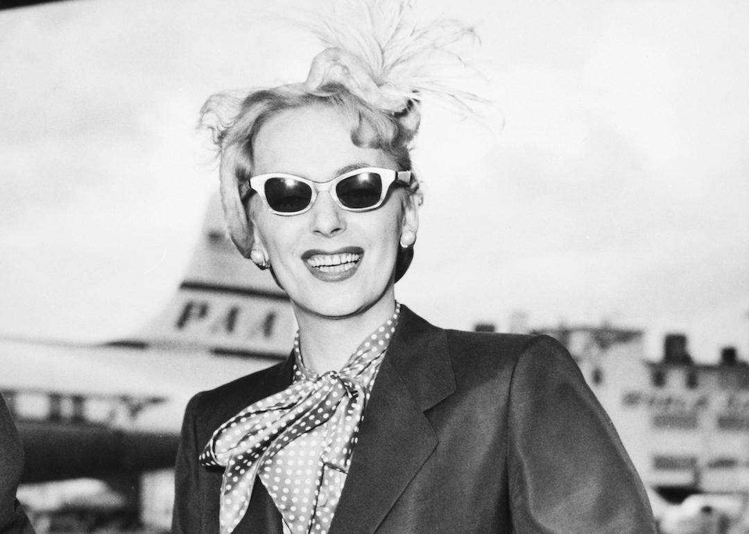 Transgender pioneer Christine Jorgensen smiles at the airport wearing sunglasses and a blazer.
