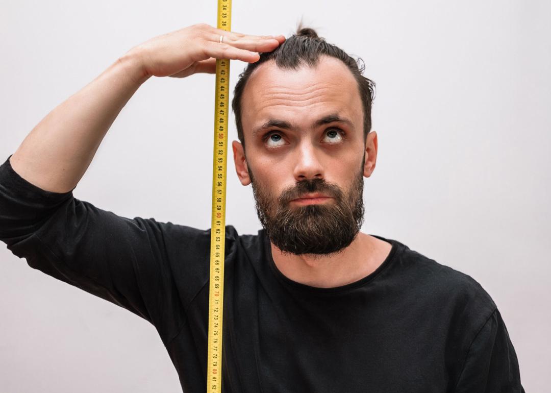 A man with a beard and a black t-shirt measures his height with a tape measure.