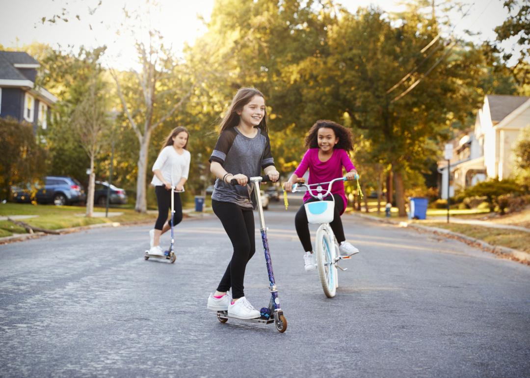 Three kids riding down the street on scooters and a bike in a picturesque suburban neighborhood.