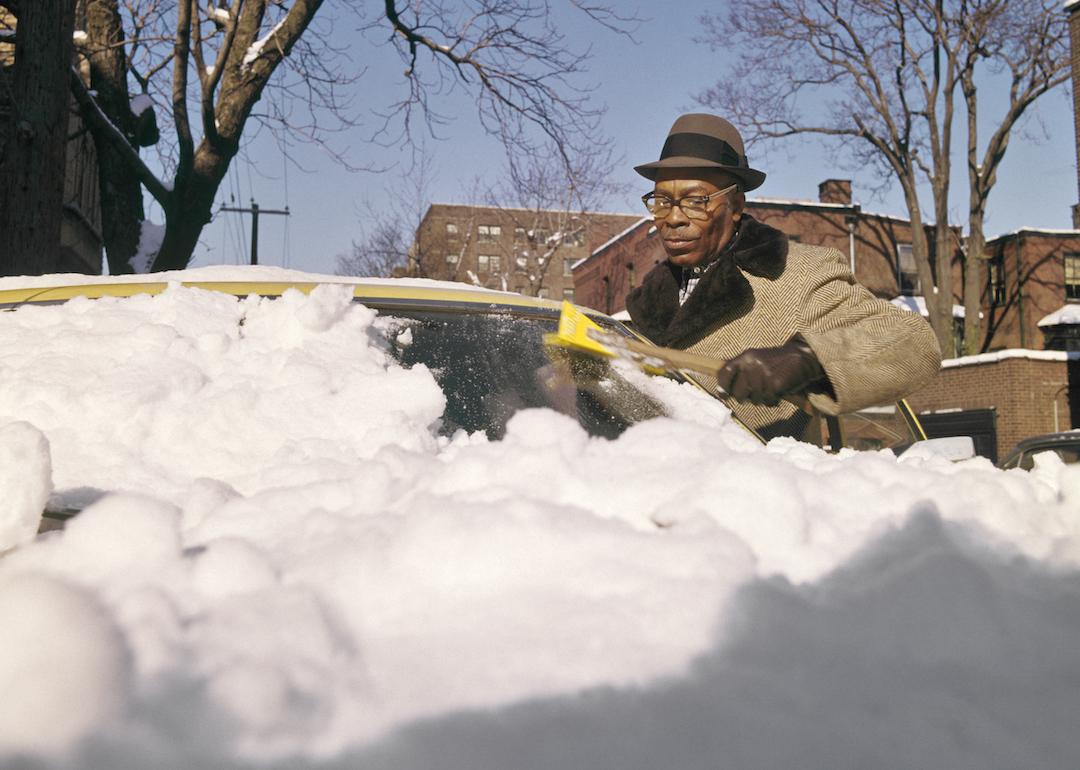A person cleans snow and ice from their car windshield in the 1970s.