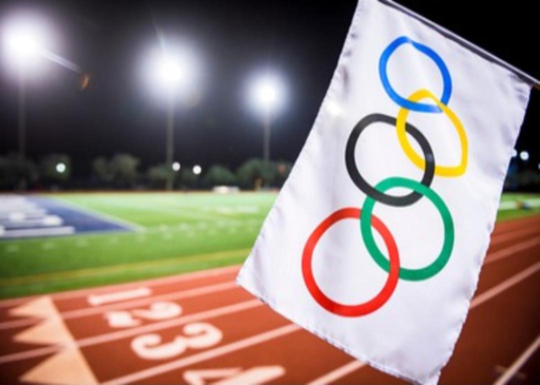 An Olympic flag waves under the floodlights of a red athletics track.