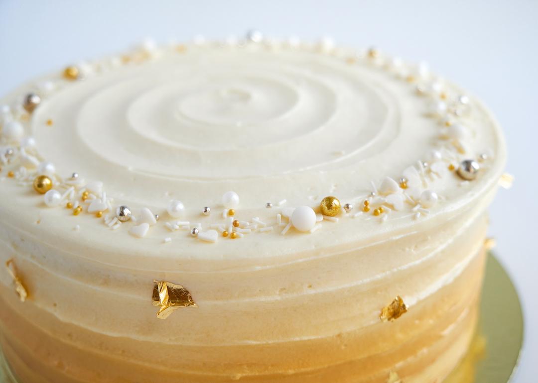 Cake with white cream, decorated with silver and gold confectionery sprinkles and gold leaf on a white background.