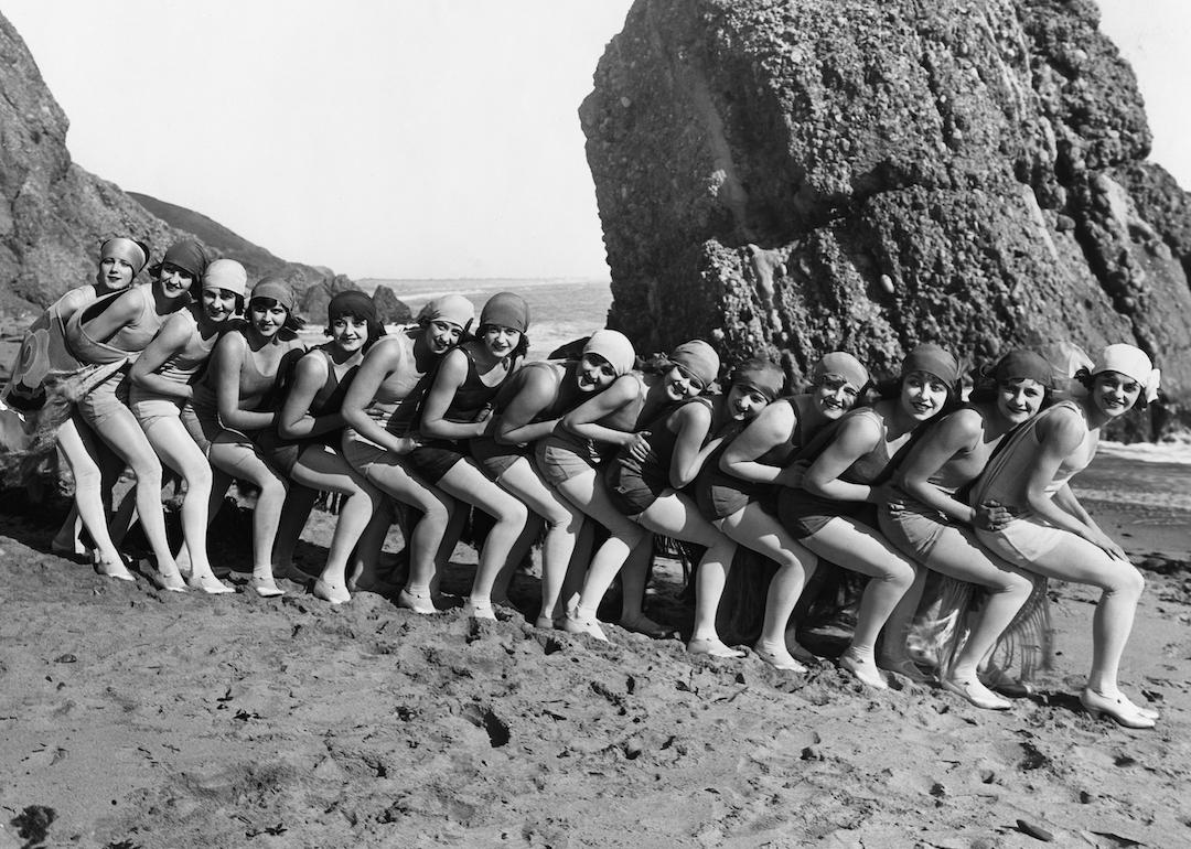 A group of young people in bathing suits and head wraps squatting in a line on the beach for a portrait, circa 1920.