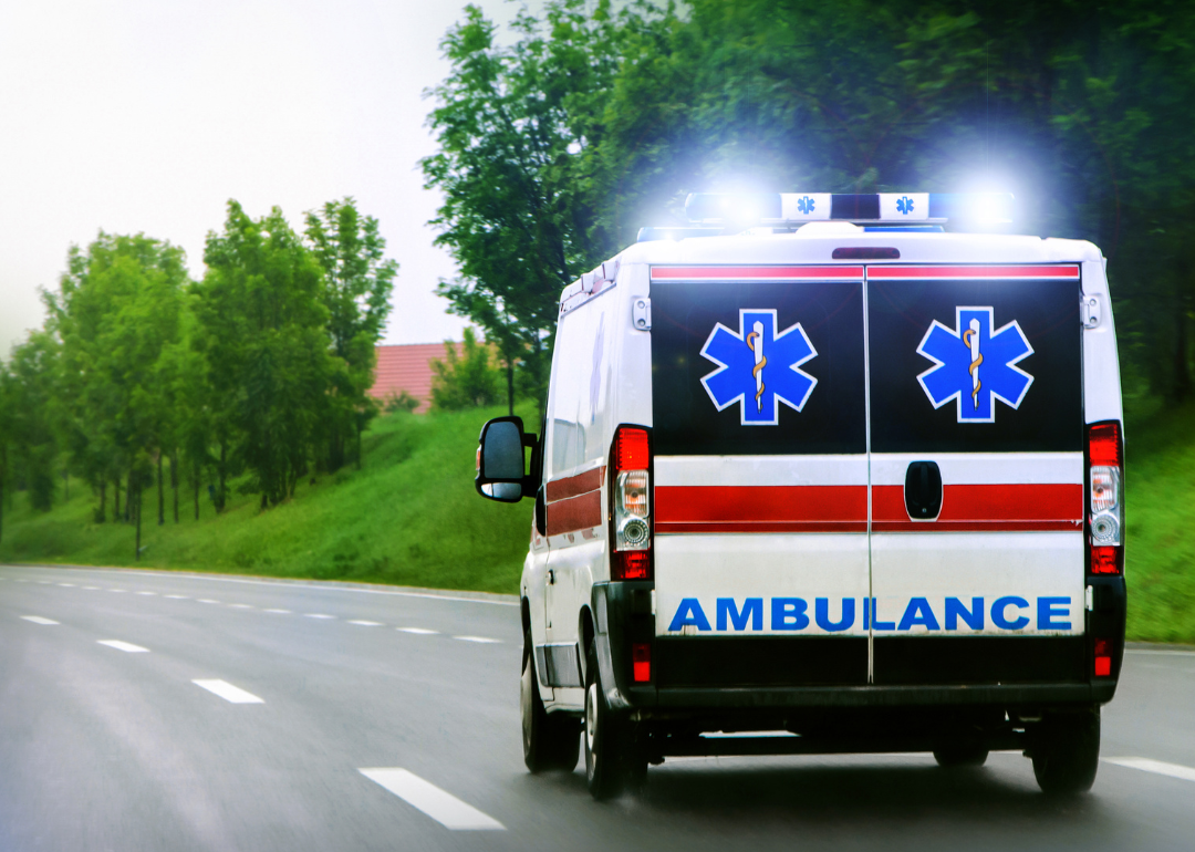 An ambulance with lights flashing drives down a road.