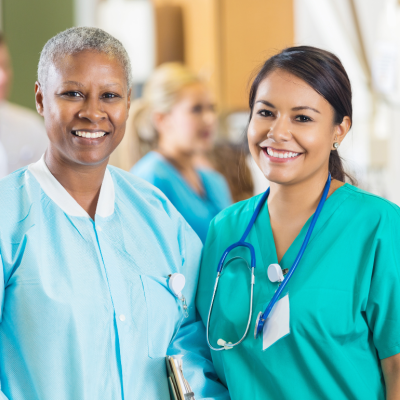 A Black and a Latina nurse in scrubs stand side by side smiling