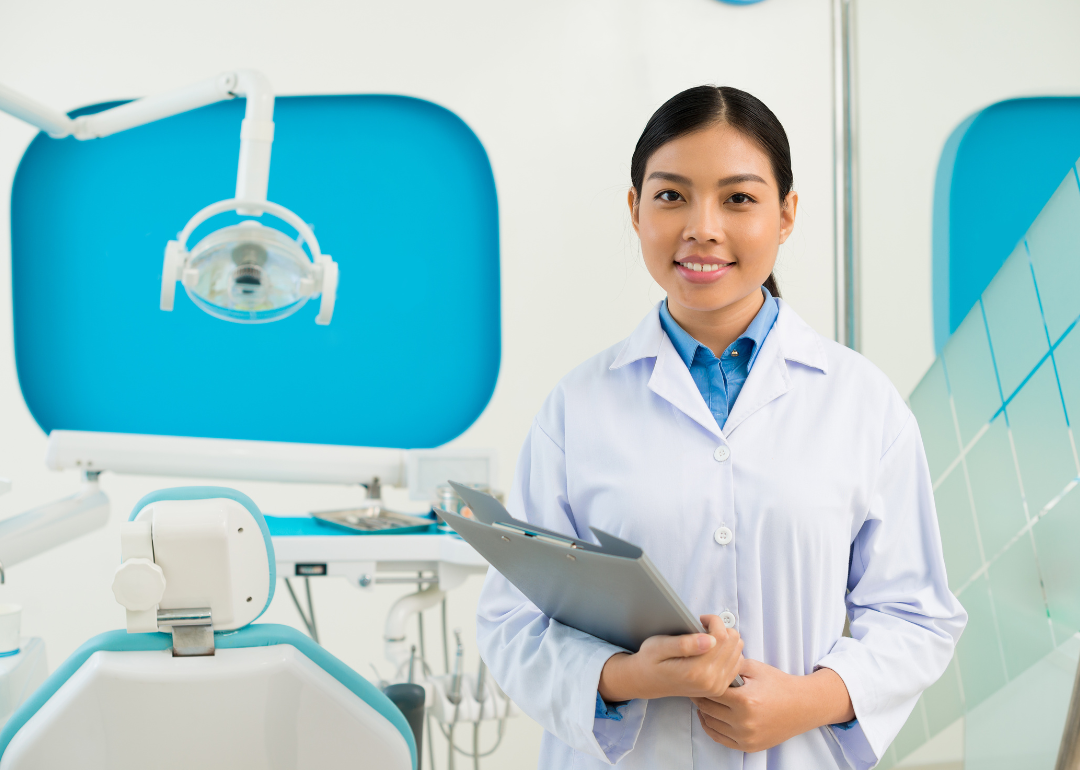 Female dental assistant standing with clipboard in front of patient chair.
