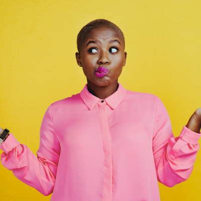 A Black woman in a pink long-sleeved blouse holds up both hands in a gesture of indecisiveness.
