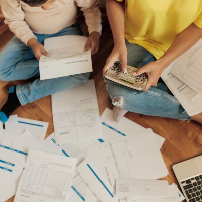 Two people sit on the floor amidst a scattering of paper bills and invoices, one of them using a calculator as they pay bills