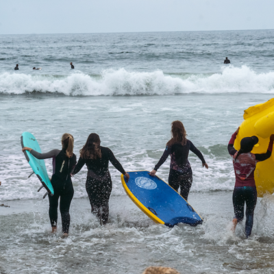 A group of women run into the ocean with body boards and an inflated floatie
