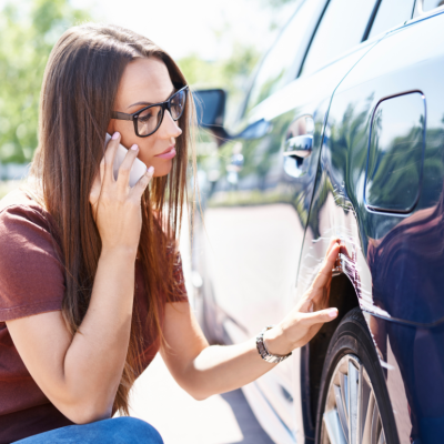 A young woman with glasses inspects damage to the side of her car and makes a phone call