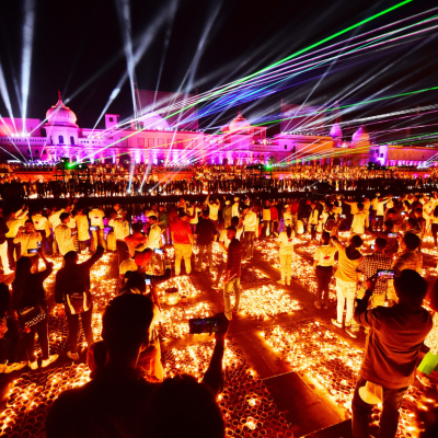 A large crowd of people stand in front of a palace with many light beams shining upward, surrounded by candles on the ground.